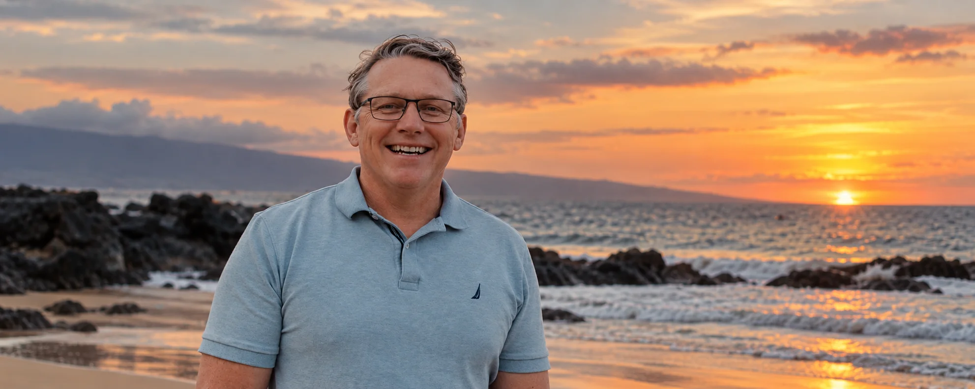 Hans Anderssen smiling on a rocky beach at sunset, the sun setting over the ocean behind him